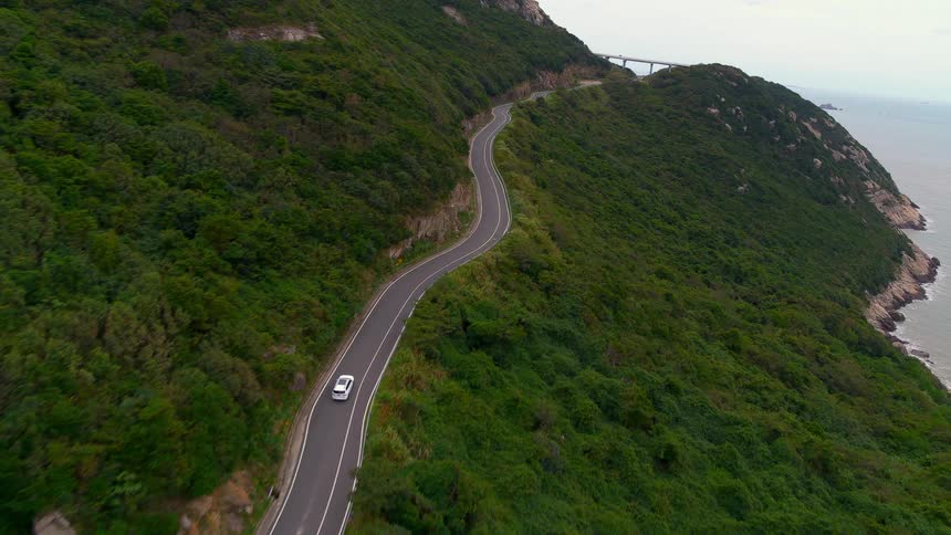 A vehicle drives on a curvy road on a hillside along the coast