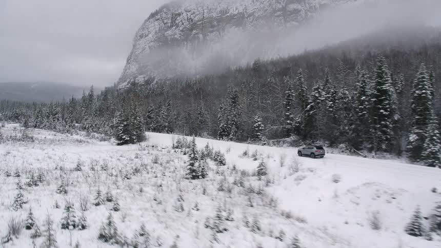 A silver car travels on a snow covered road lined with trees and a mountain