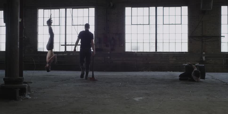 A man drags an ax into a room where one man hangs upside down and another struggles on the ground while tied to a chair