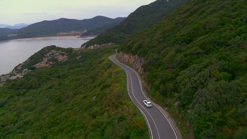 White vehicle drives along winding road hugging slope paralleling water
