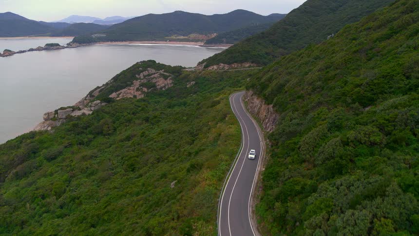 Aerial view of white car driving on highway though mountain forest next to lake