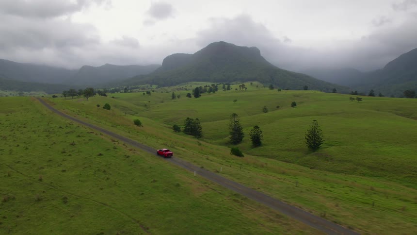 Red vehicle drives along road past green hills with fog covered mountains in distance