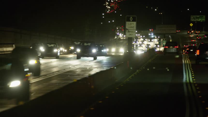 A highway with vehicles signs and streetlights with buildings in the background