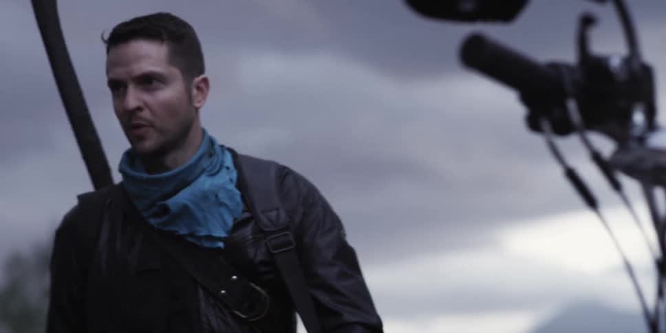 Man stands outdoors looking around near a motorcycle under overcast sky