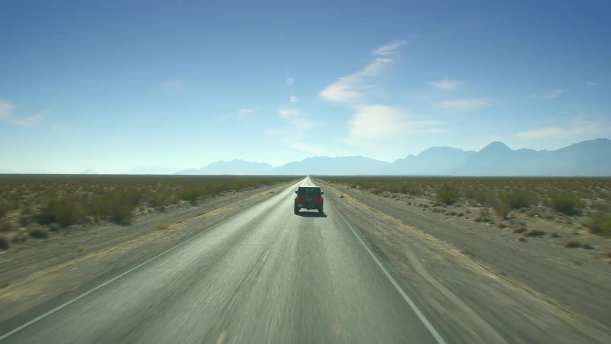 A vehicle travels down a road through a vast grassland towards distance mountains on a sunny day