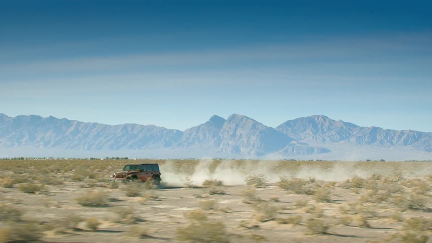 A truck is traveling across the desert leaving a trail of smoke behind it and there is a mountain range in the distance