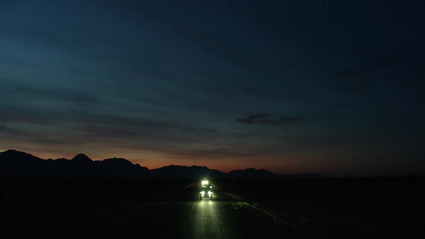 Vehicles travel along a road through a vast flatland with their headlights on at night