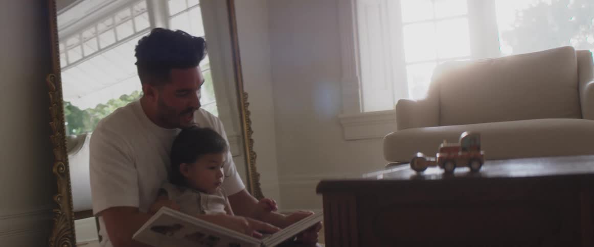A man in a room holding a child and reading  a book with a mirror a wall and window in the background