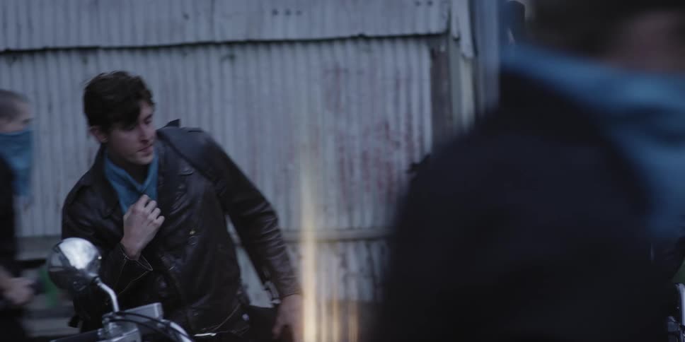Men wearing black leather jackets and blue bandanna straddle motorcycles near rusted corrugated metal sided building