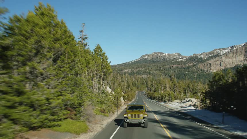 A green jeep travels on a road surrounded by trees and a mountain range is in the distance