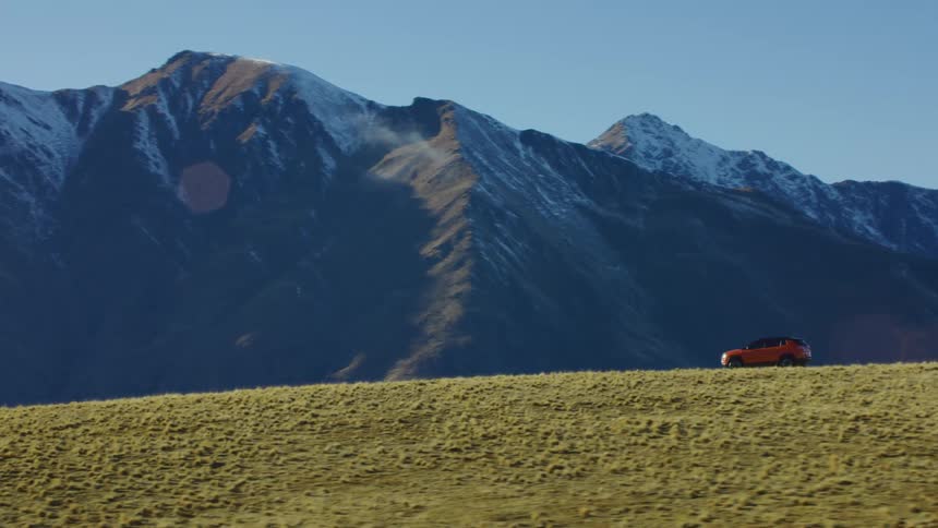 Red vehicle drives across green and tan plain with snowy mountains in background
