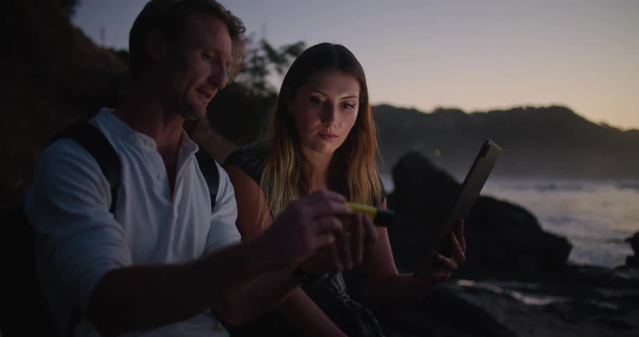 Couple looking at data on a tablet sitting near the beach at sunset