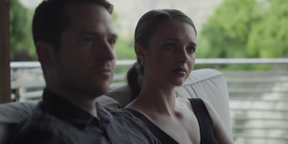 Man with stubble beard dark shirt and woman with sleeveless black shirt sit on beige couch listening to speaker and nodding