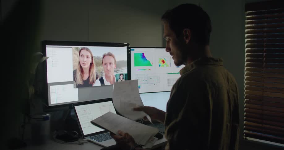 A man in a room video chatting on a computer with a window and a wall in the background