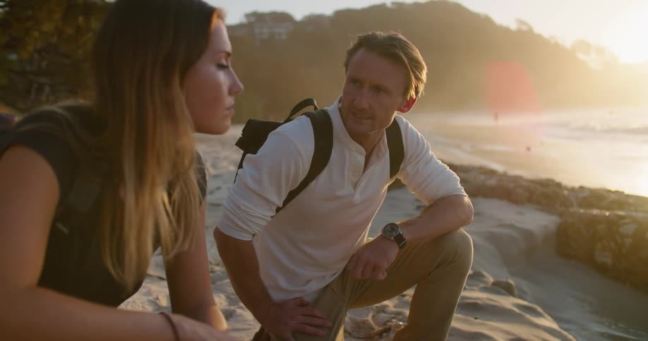 A man and woman speak with one another while kneeling on a beach just out of reach of the tide during the day