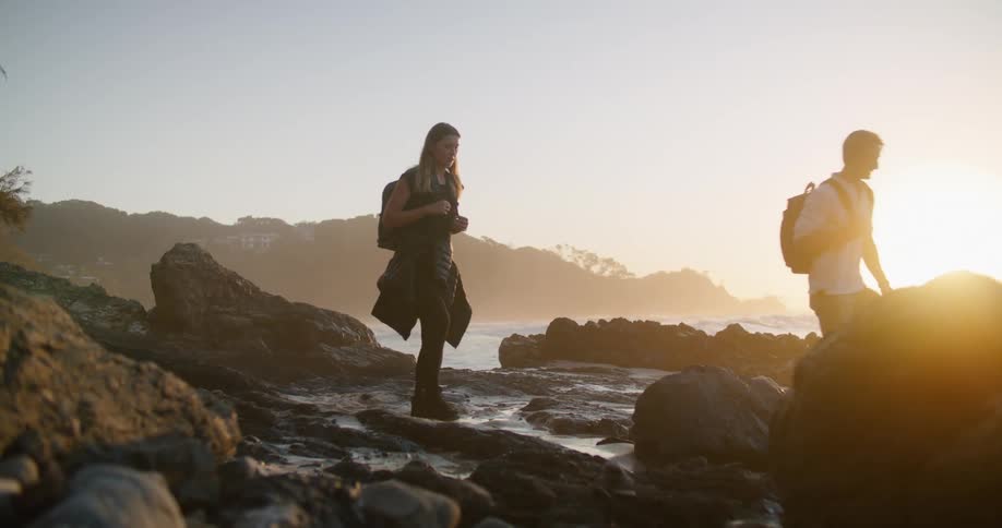 Man and woman with backpacks walk on stony beach at sunrise
