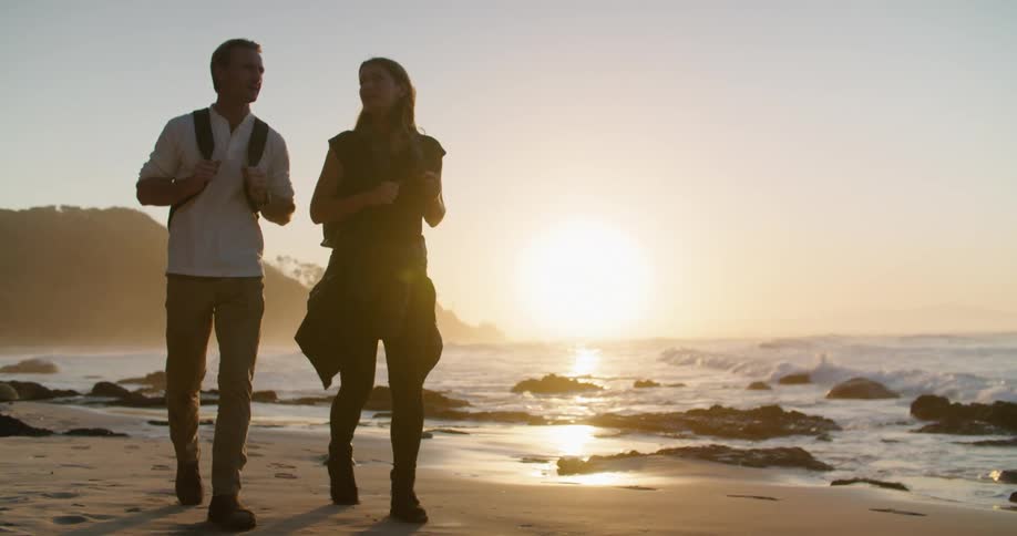 A man and woman walk across a beach as the waves roll in and a bright sunset is in the distance