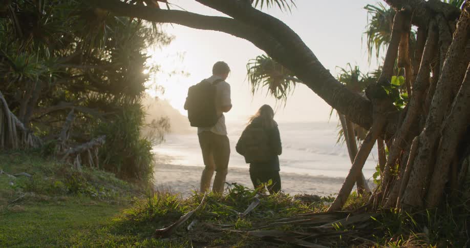 A man and woman carrying backpacks walk towards a beach as waves roll in