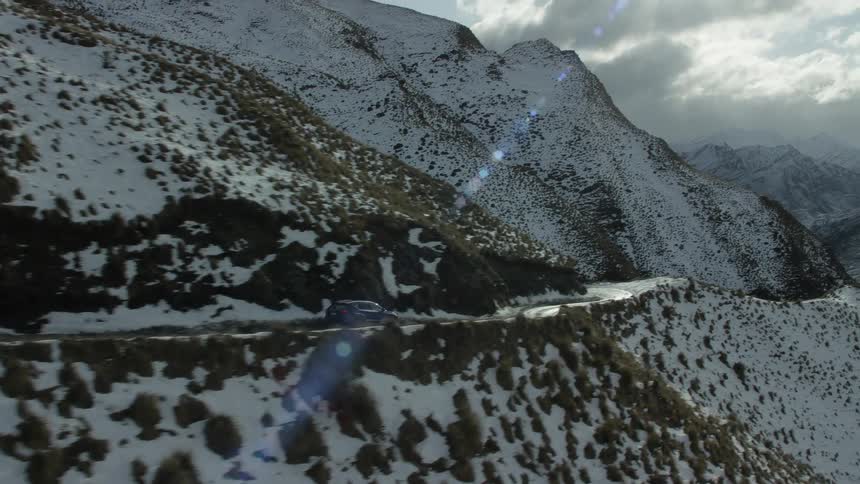 An suv travels on a narrow mountain road with slopes dotted with snow and brush