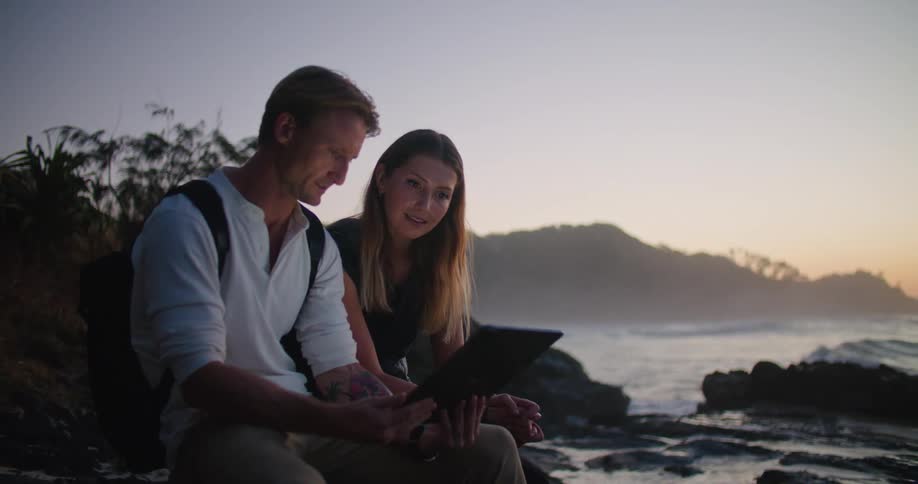 A couple sit on a rock along the coast while looking through their tablet