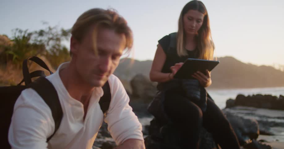 A man is testing water as a woman logs into a tablet while they sit on a rocky beach