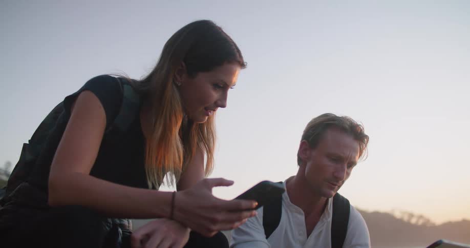 A male and female sitting near water are using a tablet and a phone