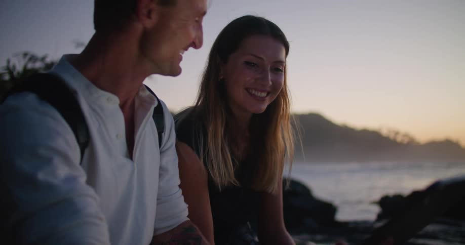 A man and woman are laughing as they sit on some rocks near the water