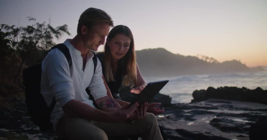 A man and woman are sitting on a rocky beach looking at a tablet when she points to the distance