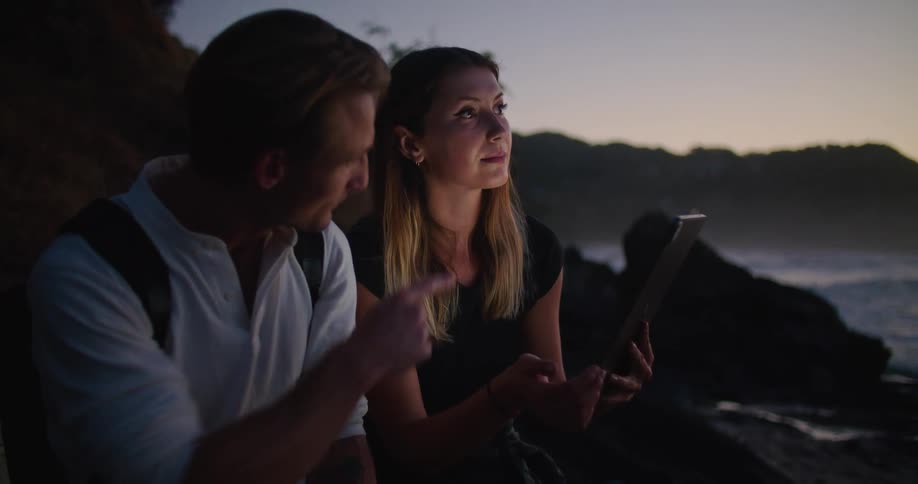 A woman and a man looking at a tablet on a beach with the sky and clouds in the background