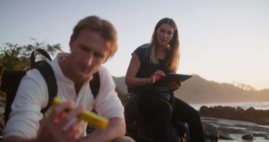 A man and woman are sitting on a rocky beach looking at an instrument and logging on a tablet
