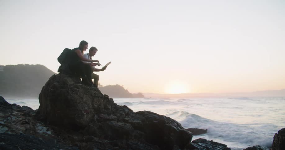 A man and a woman sitting on a rock on a beach with the ocean sky and clouds in the background