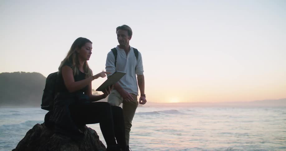 A woman uses a computer tablet while speaking to a man standing next to her on the ocean coast at sunset