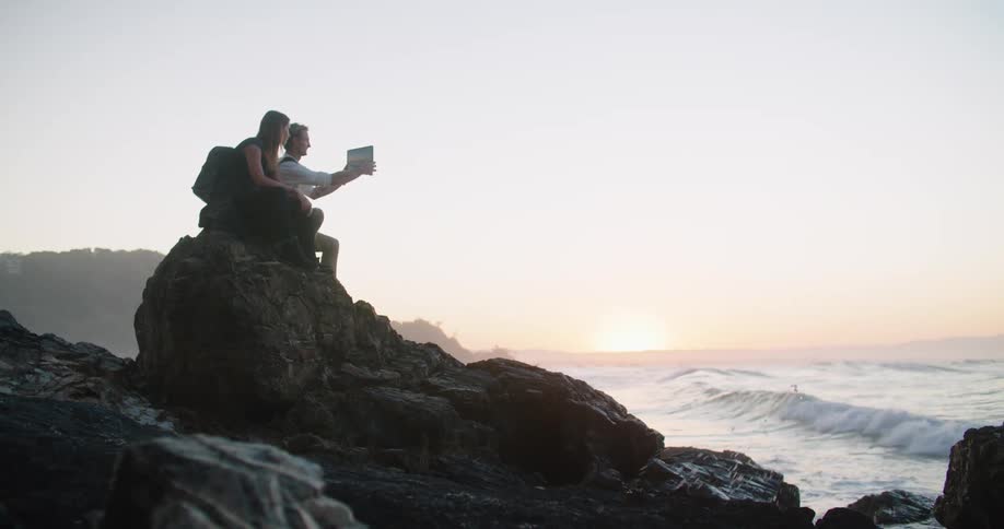 Man with white shirt backpack and woman with brown hair and backpack sitting on big rock and takes photos on tablet on the rocky beach at sunrise