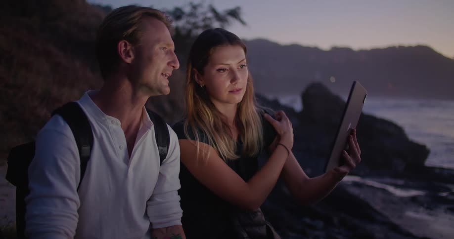 A woman in a man on a beach looking at a tablet with the sky and clouds in the background