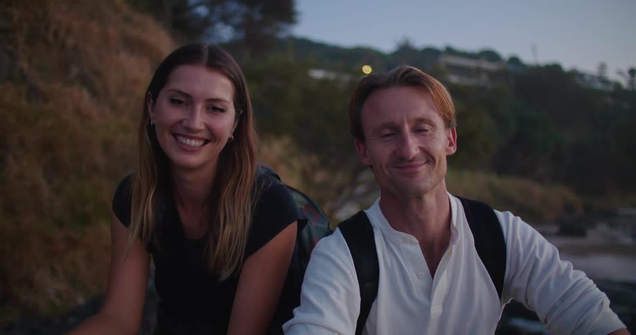 Young couple sitting near the beach at sunset looking at the camera