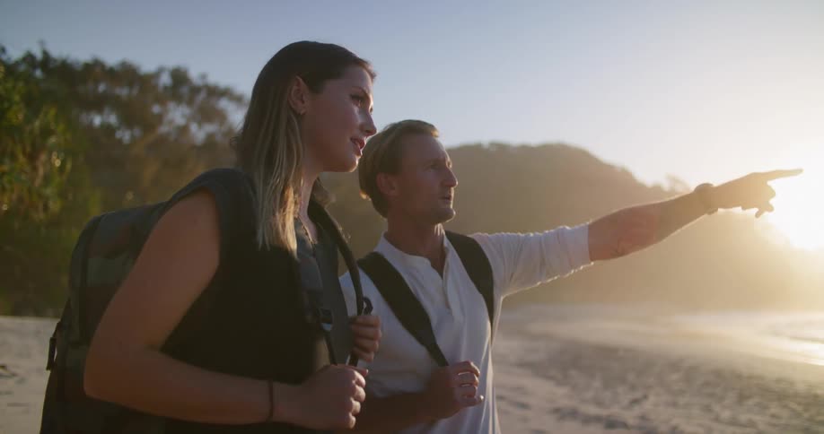 Two people with backpacks stand on the beach at sunset and point to the sea