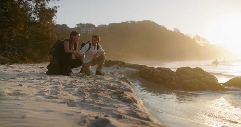 A man and woman speak to one another while kneeling side by side on a beach and looking at the tide