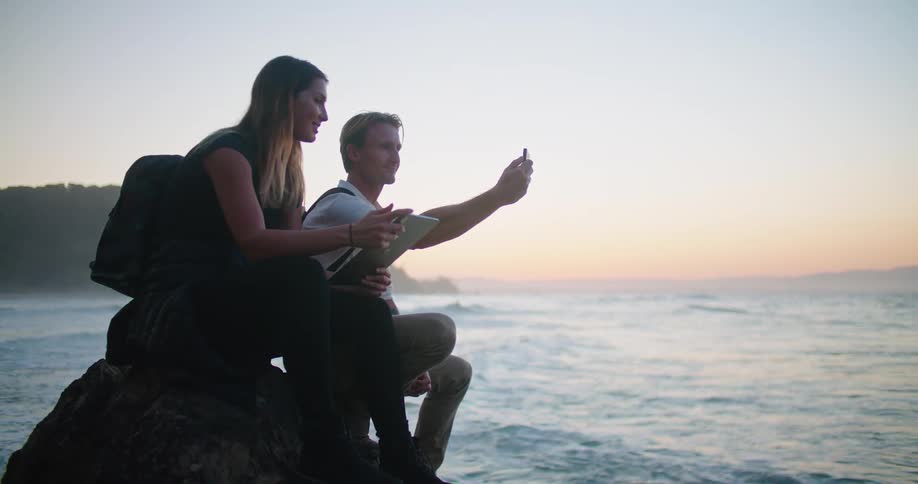 A man and woman sitting on a rock having a discussion while filming and tapping on a tablet