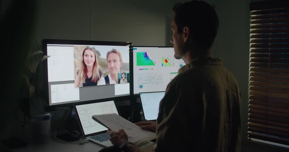 A man at a desk with two laptops and two monitors picks up some papers and talks to two people he is video chatting with