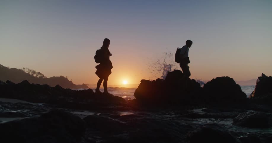 Silhouette of two people wearing backpacks walking on rocks along the coast as waves roll in and a bright sunset in the distance