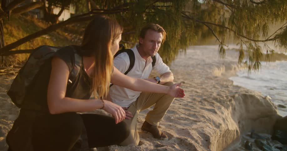 Young couple kneeling under a tree near the coast close to the ocean at sunset