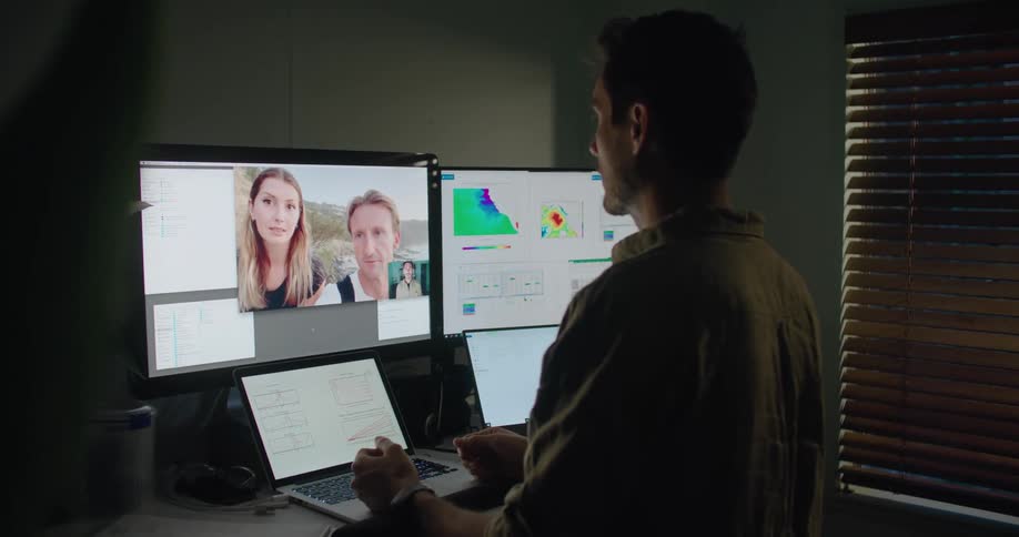 Man standing in dimly lit office at table with several computers and screens talks via video conferencing with man and woman on beach