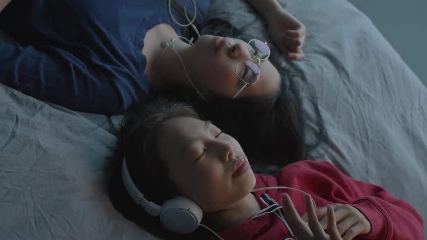 Two girls with red zip up and dark blue shirt lie on bed and listen to music through headphones and earphones in room
