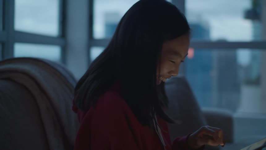 A girl laughs as she plays a video game on a tablet sitting on a couch near large windows