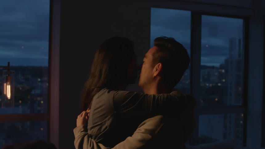 Man cuddles and kisses woman in dim lit apartment room next to windows