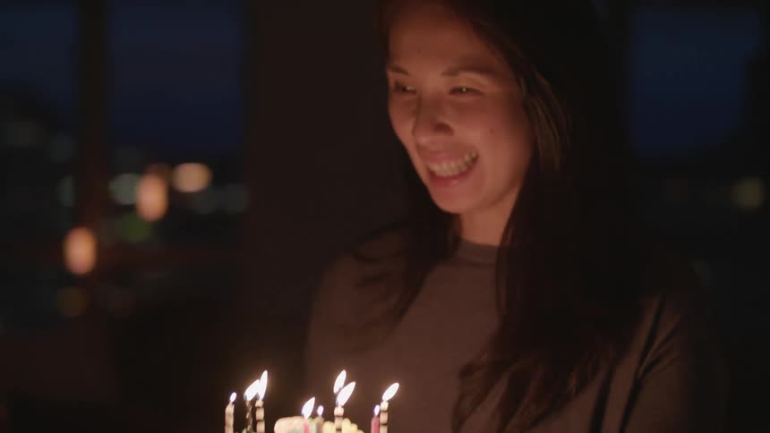 A woman smiles as she brings a birthday cake to a table surrounded by other people