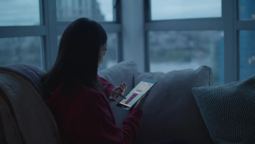 Woman sits on couch near windows showing city while scrolling on tablet