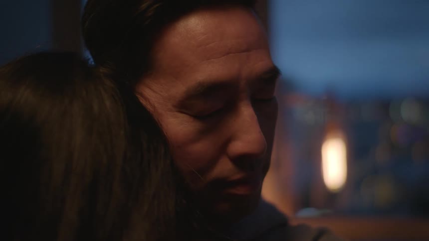 Father cuddles and kisses girl wearing grey hoodie in dim lit apartment room