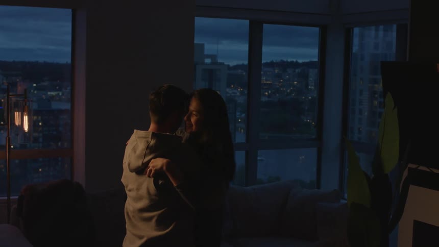 A couple dance in each others arms while smiling in an apartment overlooking a city skyline