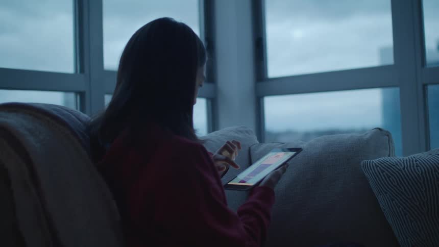 Girl with straight hair and sweater sits on grey couch and touches tablet screen in living room at home